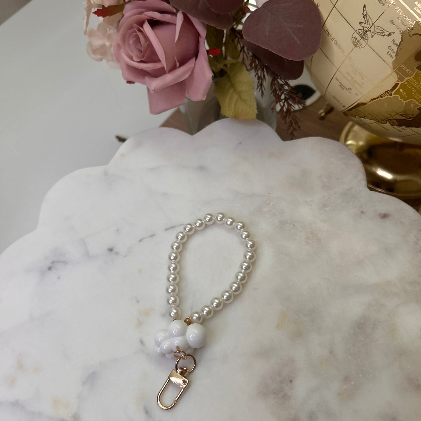 Pearl bracelet with a gold clasp on a marble surface next to pink flowers and a globe.