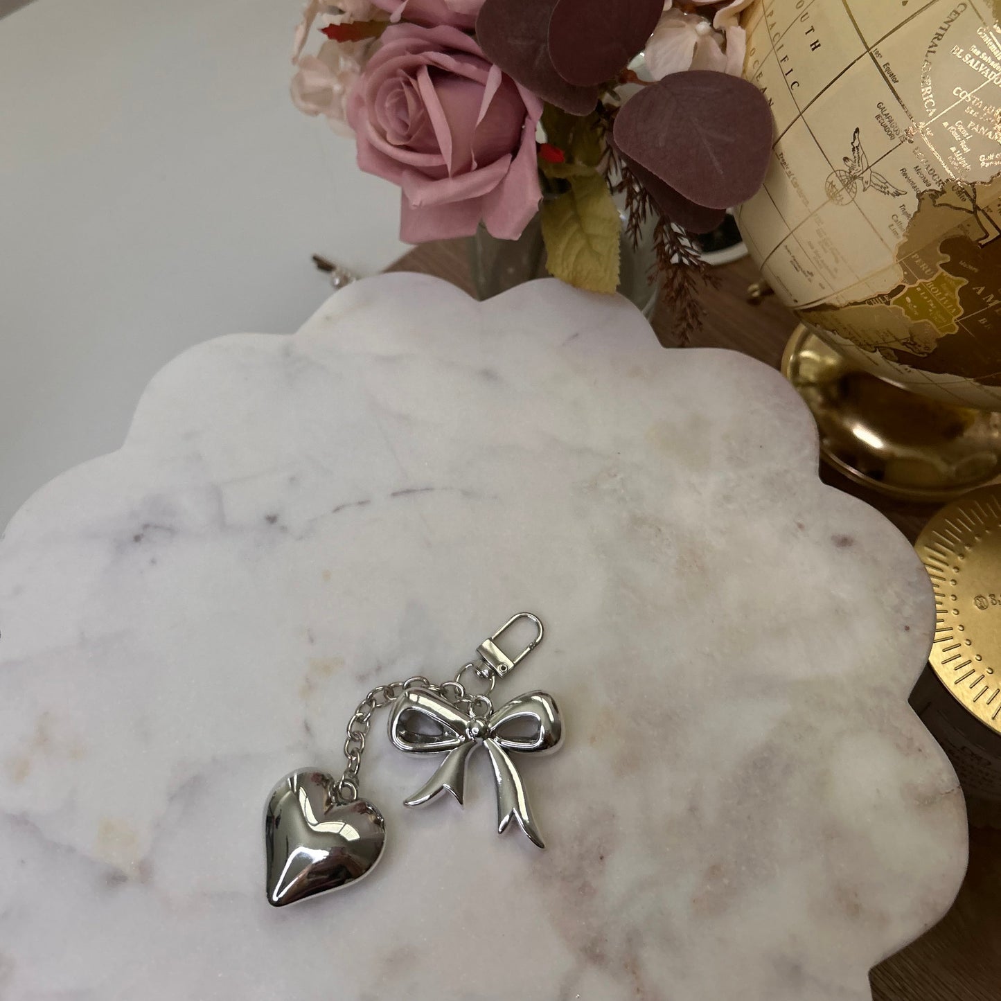 Silver keychain with heart and bow design on a marble surface with flowers and a world map in the background.